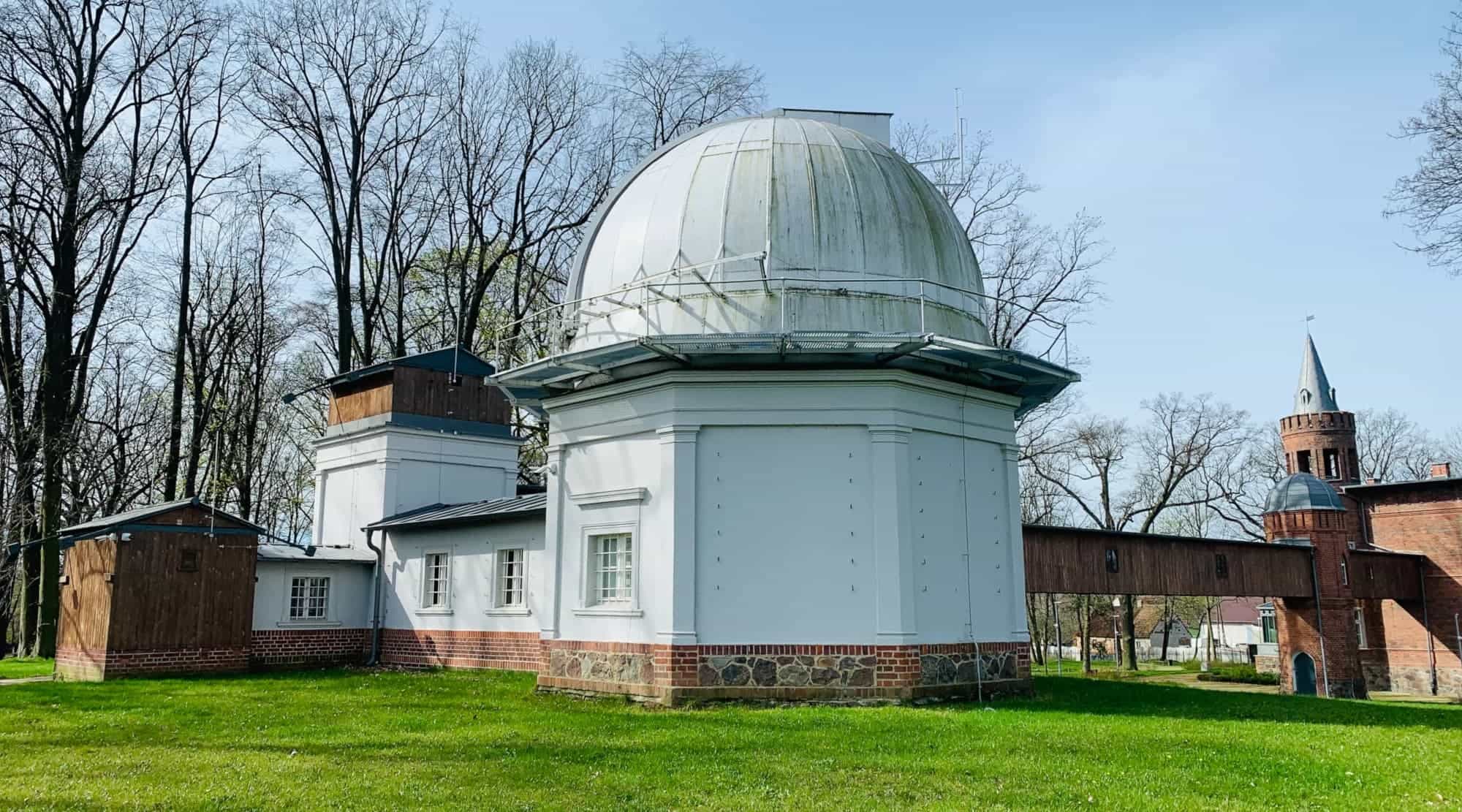 White observatory dome and historic observatory building surrounded by grass and trees in daylight.
