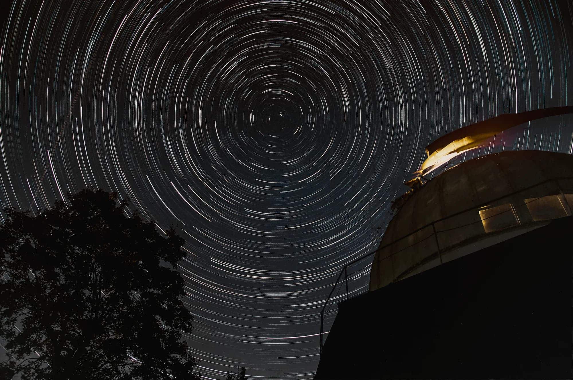 Long-exposure night photograph showing circular star trails above an observatory dome and nearby trees.